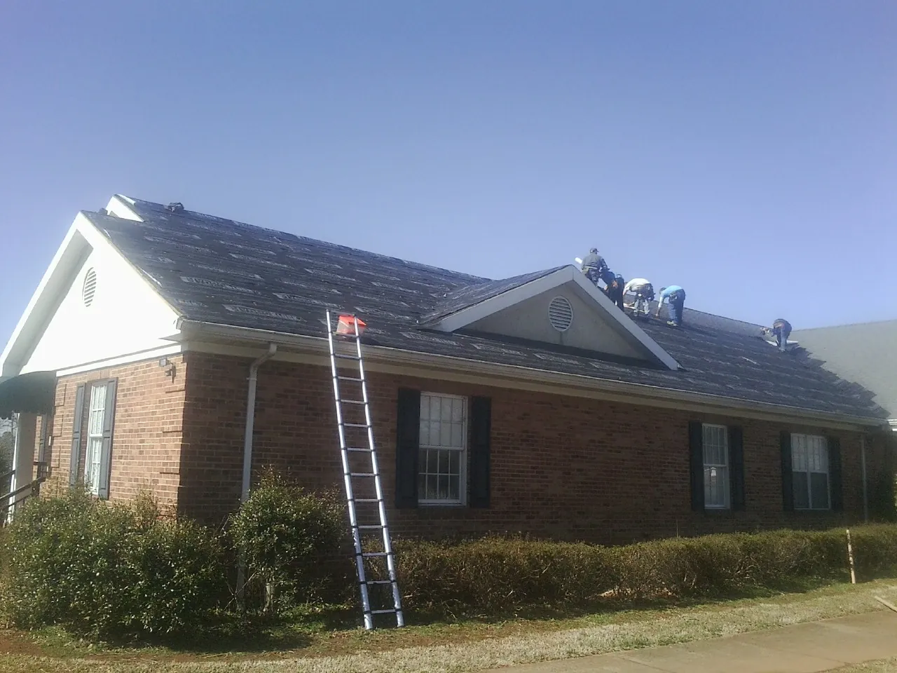 Crew tearing shingles off a brick ranch roof with a ladder set — Metal Roof Repair work by our crew in Amity